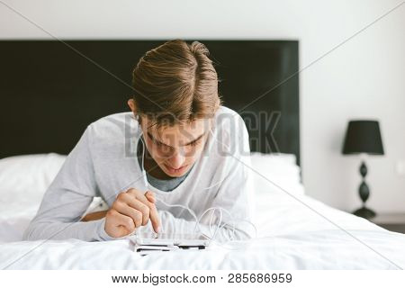 16 years old teenager using tablet pc and listening to music in earphones while relaxing on couch in his room