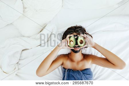 Lifestyle image of 12-14 years old teen girl applying black clay facial mask with avocado. Top view photo of teenage girl wrapped in towel doing organic anti blemish skin care routine.