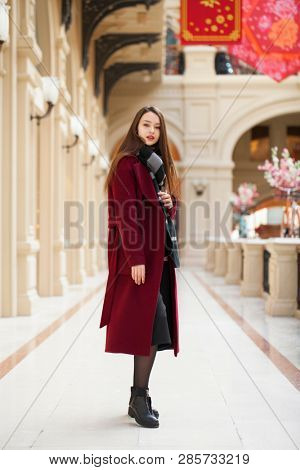 Full body portrait. Young beautiful woman in stylish in a long burgundy coat, indoor shop