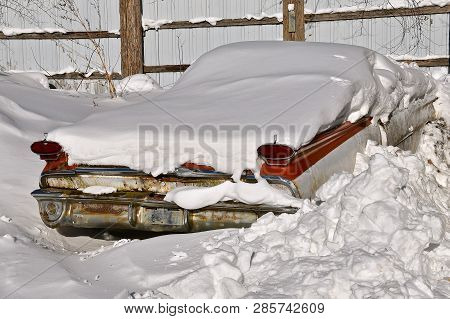An Old Rusty Car Is Buried In A Snowdrift With Only Blinkers, Taillights And Bumper Exposed.