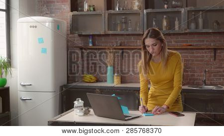 Businesswoman Writing Notes In Kitchen. Adult Woman Working At Home On The Cooking Table Laptop. Cau