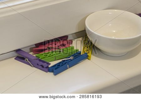 Colored Clothespins And A White Bowl Next To Them On The White Windiw Sill Background. Hanging And D