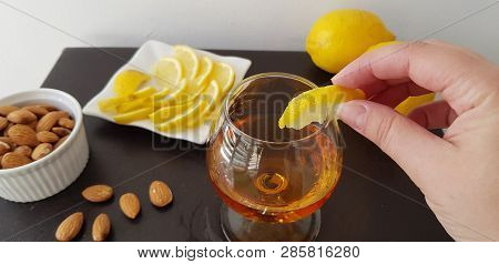 Female Puts A Slice Of Lemon On Glass Half Full With Brandy On Brown Table Near Roasted Almonds And 