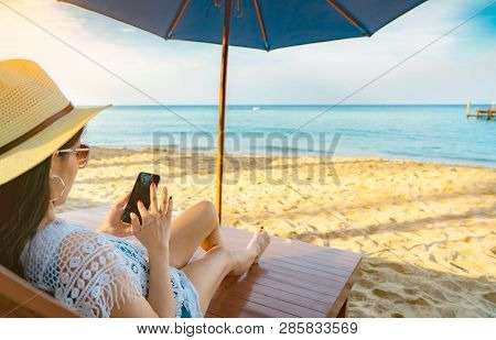 Asian Woman With Hat Sit On Sunbed Under Beach Umbrella At Sand Beach And Using Smartphone On Summer