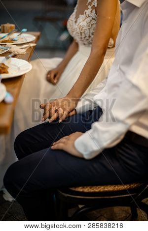 A Groom And A Bride Are Holding Hands. They Are Sitting Behind The Table.