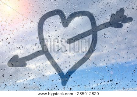 Figure Heart Pierced By Cupid, On The Misted Glass. Glass Window With Raindrops Against The Sky. Unr