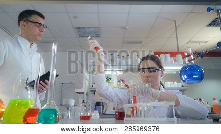 Woman Adding Liquid To Test Tubes With A Micropipette. Two Olleagues Performs Medical Tests In Micro