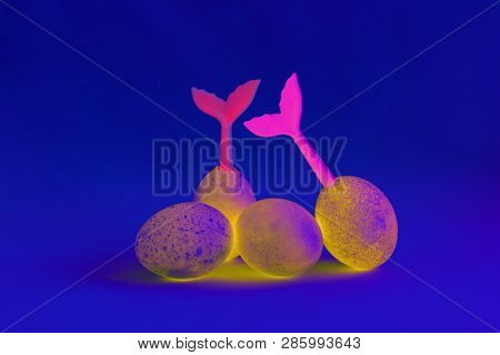 Four Yellow And Magenta Easter Eggs Lie On A Blue Background. Tails Of Mermaids Stick Out Of Eggs. S