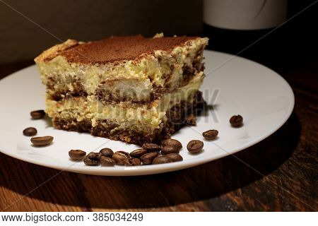 Slice Of Gourmet Tiramisu Surrounded By A Sprinkling Of Coffee Beans On A Thin White Plate Atop A Ru