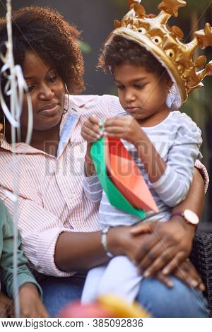 little afro-american girl sitting in the lap of her mom with golden yellow inflatable crown on her hed, playing with paper boats 