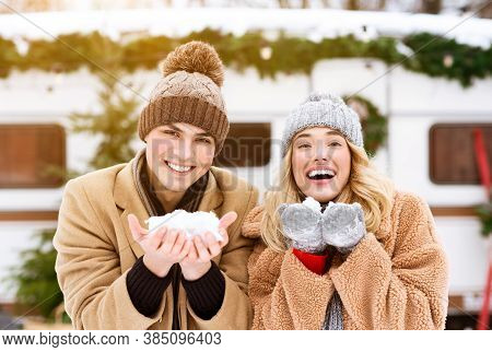 Winter Mood. Cheerful Young Couple Holding Snow In Hands And Smiling At Camera, Standing At Campside
