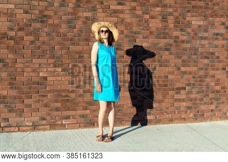 Slender Caucasian Woman In Blue Dress And Straw Hat With Fatty Shadow On The Wall. The Concept Of In