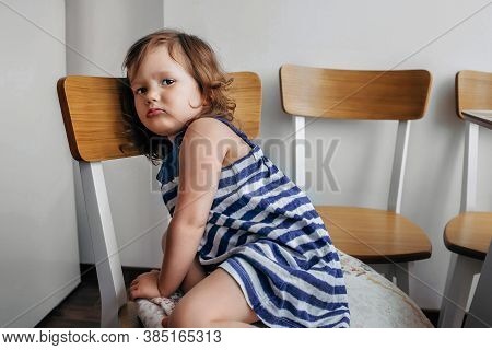 Child Girl Sitting On A Chair And Looking Sadly At The Camera.