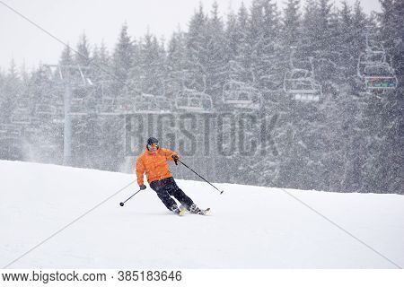 Adult Proficient Man Skiing On Ski Run Slope During Heavy Snowfall. Chairlift And Fir Forest On Back