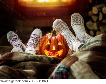 Female and childish feet  are in soft wool socks with burning fireplace at the background. Halloween celebration. Carving jack-o-lantern pumpkin is on the table.