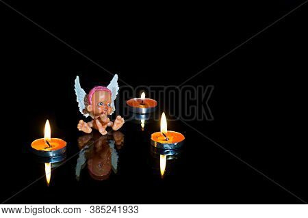 The Colored Little Toy Angel And Candles In The Darkness, Reflection, Black Background. Mysticism