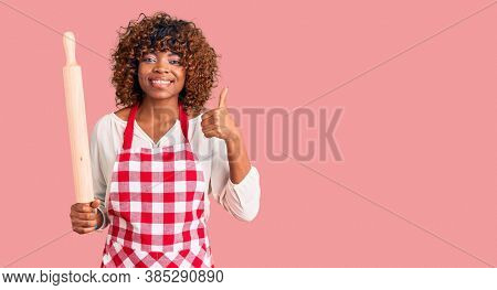 Young african american woman wearing apron holding rolling pin smiling happy and positive, thumb up doing excellent and approval sign 