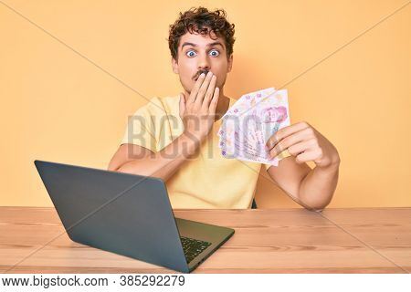 Young caucasian man with curly hair sitting on the table working with laptop and holding mexican pesos banknotes covering mouth with hand, shocked and afraid for mistake. surprised expression 