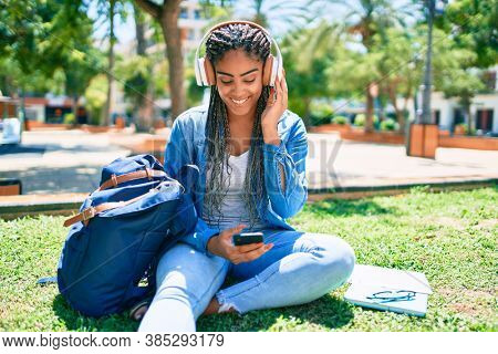 Young african american student woman smiling happy listening to music sitting on the grass at the university campus