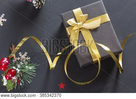 Christmas Gift Box With A Gold Bow On A Black Background. Spruce Branch With Red Berries. Copyspace