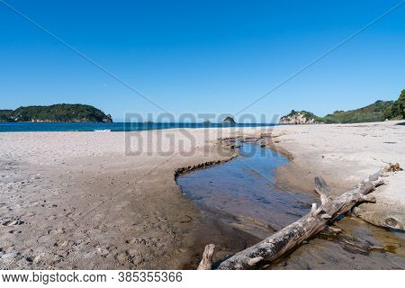 Hahei Beach White Sand And Blue Sea With Small Stream Through Sand And Driftwood Log Laying Across.