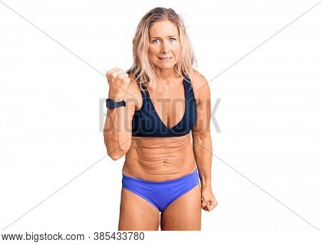 Middle age fit blonde woman wearing bikini angry and mad raising fist frustrated and furious while shouting with anger. rage and aggressive concept. 