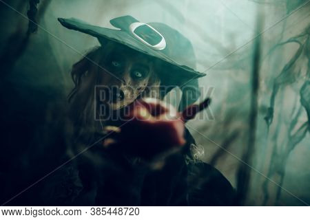 Halloween. Scary old witch holds out a red apple. Woodland background, evil black magic.