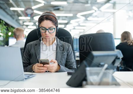 Business Woman Dressed In A Headset Is Bored And Uses A Smartphone While Sitting At A Desk. Female M