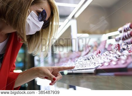 Young Woman In Medical Mask, Sunglasses, Red Jacket Points Her Finger At Jewelry Display. Social Dis
