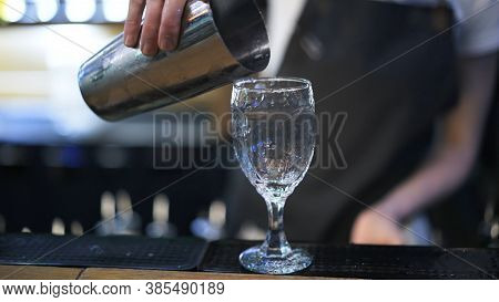 Holding Metal Cup To Pour Cocktail Into A Glass, Background Of Bartender. Making Cocktail In Night C