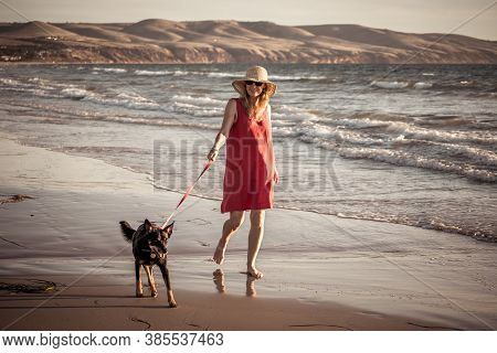 Happy Attractive Mature Woman With Her Pet Walking On Friendly Dog Beach At Sunset