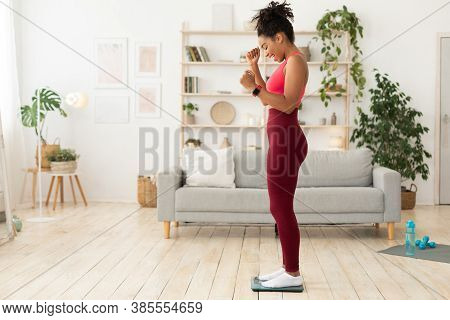 Joyful Skinny Black Woman Weighing Herself After Successful Dieting And Weight Loss Standing On Weig
