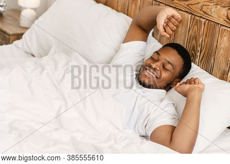 Happy Rested Wellslept Black Man Waking Up In The Morning Stretching Hands Lying In Comfortable Bed 