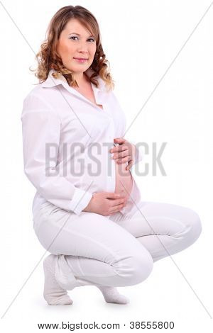 Sorridente donna incinta in bianco squat su cosce e tiene il suo ventre isolato su sfondo bianco.