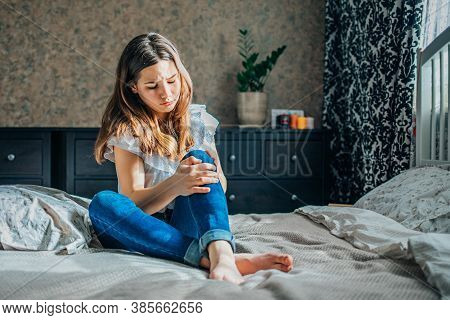 Young Brunette Girl In A White Blouse And Blue Jeans Sits On A Bed In Her Room, Clutching A Sore Kne