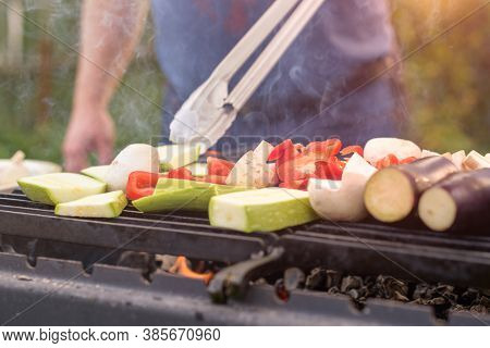 A Man Turns Over Food Tongs And Roasts Grilled Pieces Of Zucchini, Eggplants, Sweet Paper And Mashro