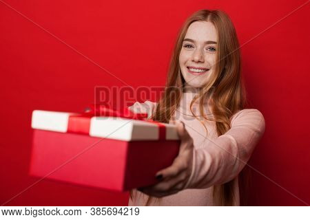 Caucasian Woman With Freckles And Ginger Hair Is Giving A Box With Gift At Camera And Smile On Red S