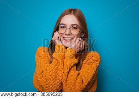 Charming Ginger Woman With Glasses And Yellow Sweater Is Gesturing Warmth On A Blue Studio Wall Smil