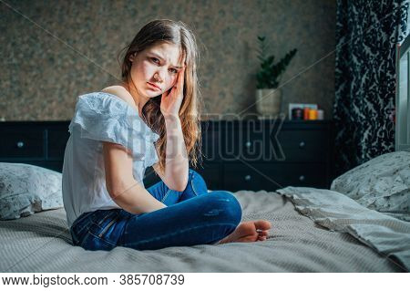A Sad Dark-haired Girl In A White Blouse And Blue Jeans Is Sitting On The Bed In Her Bedroom. She Ha