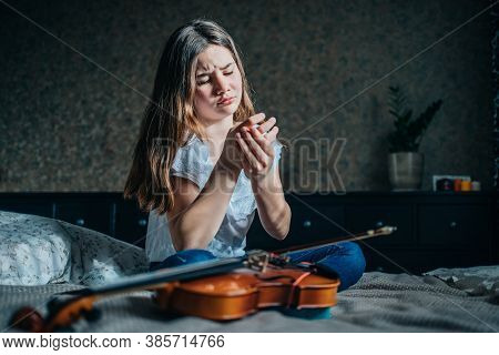 A Young Girl Sits On A Bed In Her Room, Next To Her Lies A Violin, She Holds A Wounded Finger With H