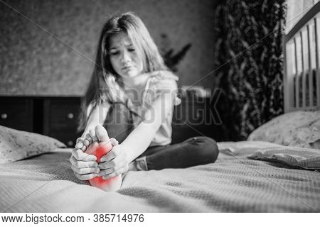 A Dark-haired Girl In A White Blouse And Blue Jeans Is Massaging Her Leg. She Is Sitting On The Bed 