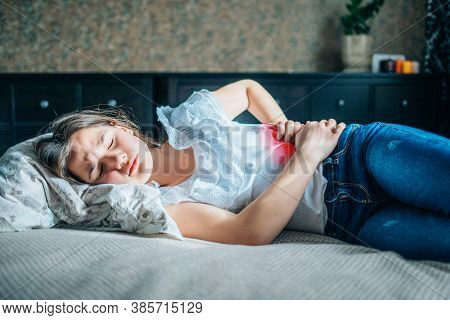 Young Brunette Girl Lies On A Bed In Her Room, Holds Her Right Side With Her Hands. Red Spot