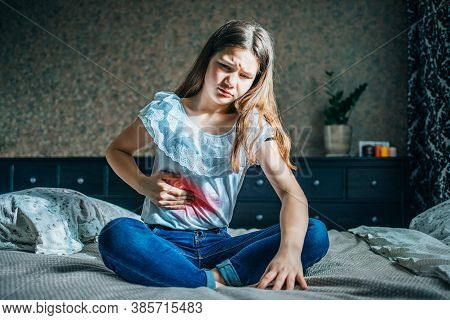 Young Brunette Sits On A Bed In Her Room, Holds A Sick Stomach With Her Hand