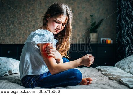 Young Brunette Girl Sitting On The Bed In Her Room, Holds An Injured Shoulder With Her Hand, Horizon