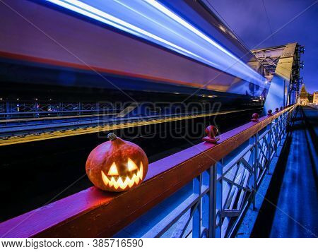 Halloween Pumpkin With A Glowing Grimace At Night On The Railway Bridge. In The Background There Are