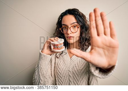 Young beautiful woman with curly hair holding plastic teeth with dental baces with open hand doing stop sign with serious and confident expression, defense gesture