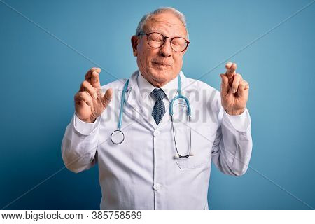 Senior grey haired doctor man wearing stethoscope and medical coat over blue background gesturing finger crossed smiling with hope and eyes closed. Luck and superstitious concept.