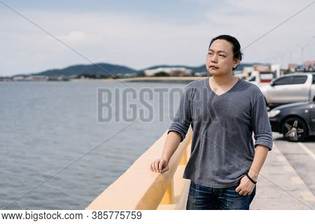 Asian Male Long Hair With Dark Gray T-shirt Stands On The Bride Beside The Road And Looks Forward To