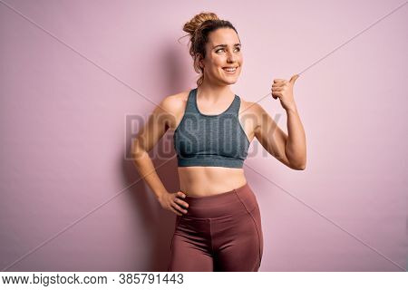 Young beautiful blonde sportswoman doing sport wearing sportswear over pink background smiling with happy face looking and pointing to the side with thumb up.