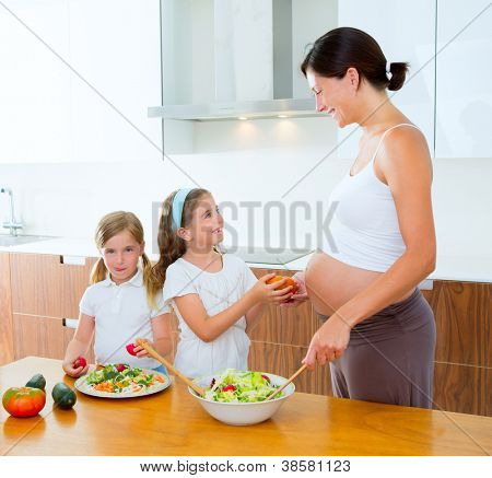 Bellissima mamma incinta con le sue figlie in cucina preparando insalata